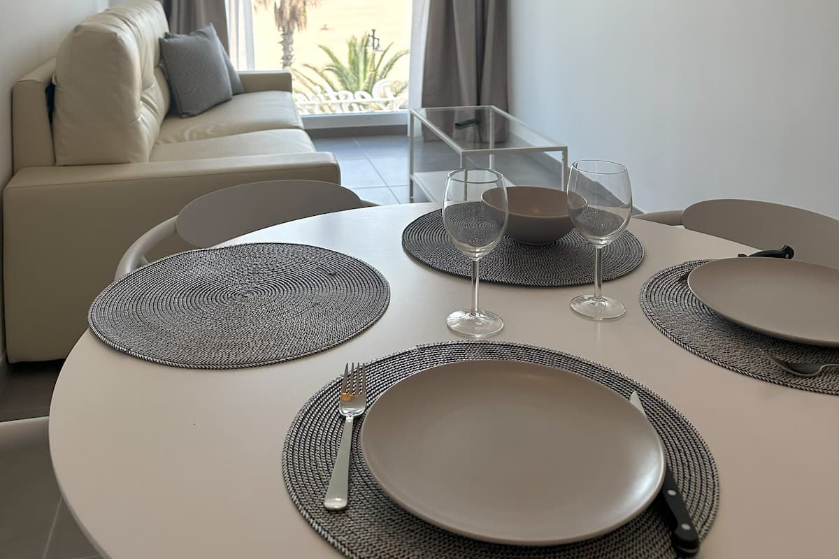 Un salon moderne avec un canapé, une table à manger pour deux personnes, un téléviseur fixé au mur et une grande fenêtre avec des rideaux.