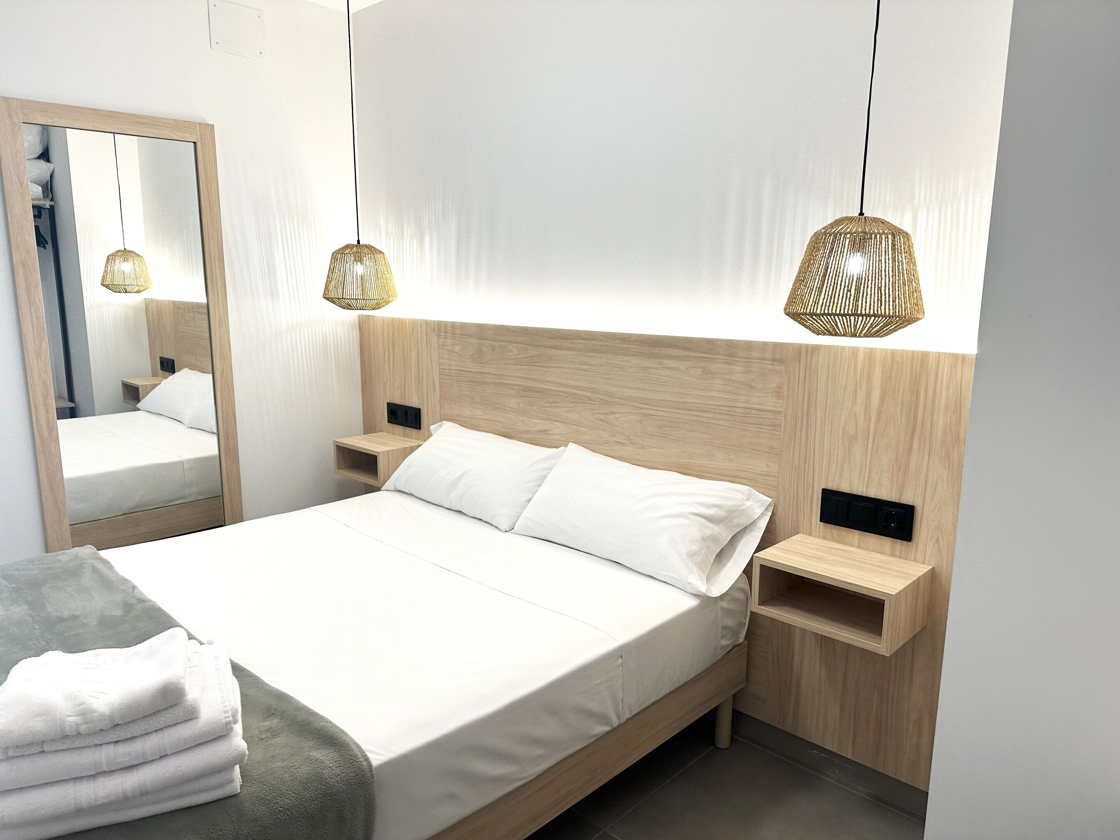 Une chambre moderne avec une tête de lit en bois, un grand miroir, des tables de chevet et deux lampes suspendues.