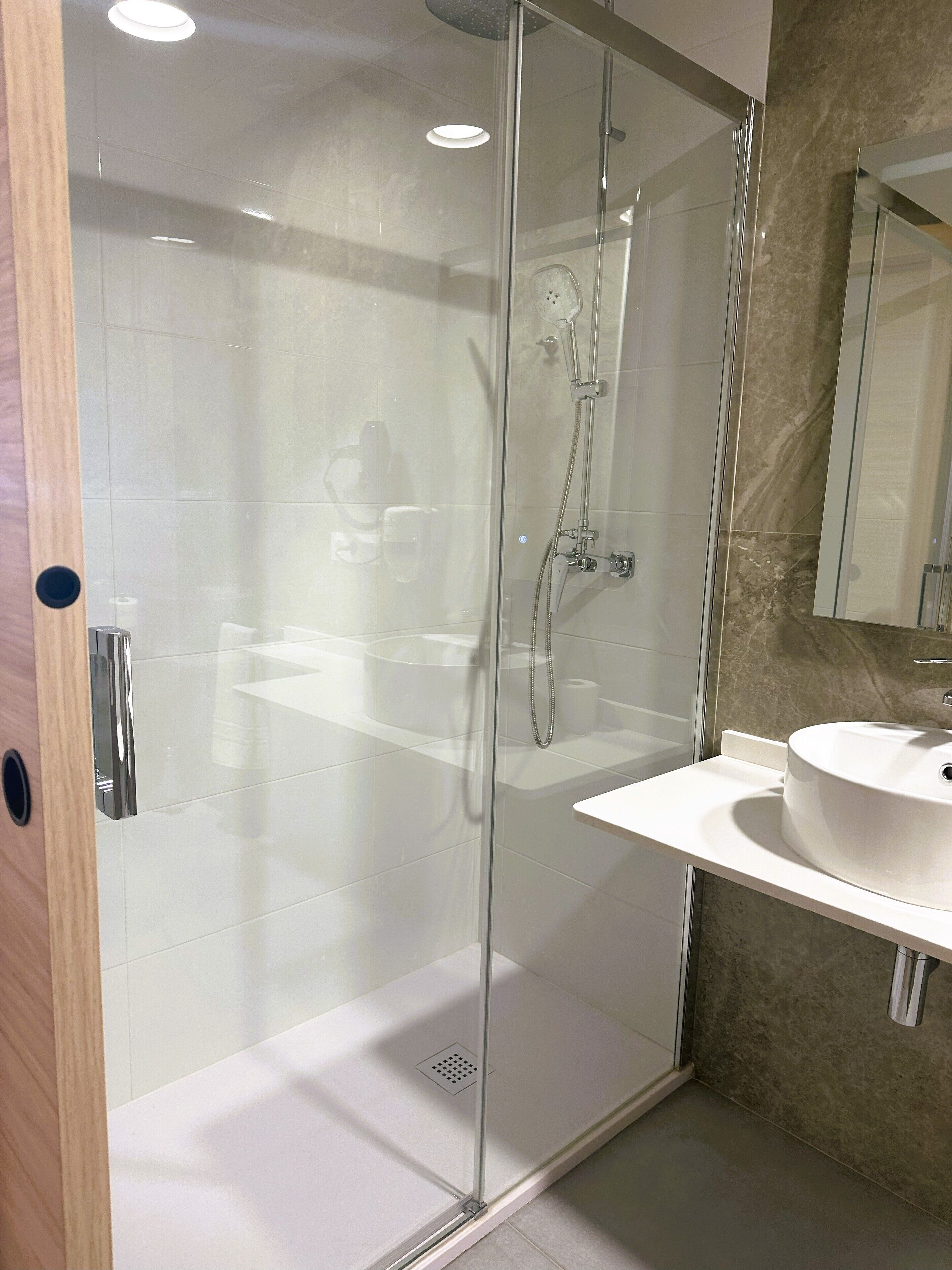 Un baño moderno con ducha envuelta en vidrio, lavamanos blanco y pared con azulejos de mármol.