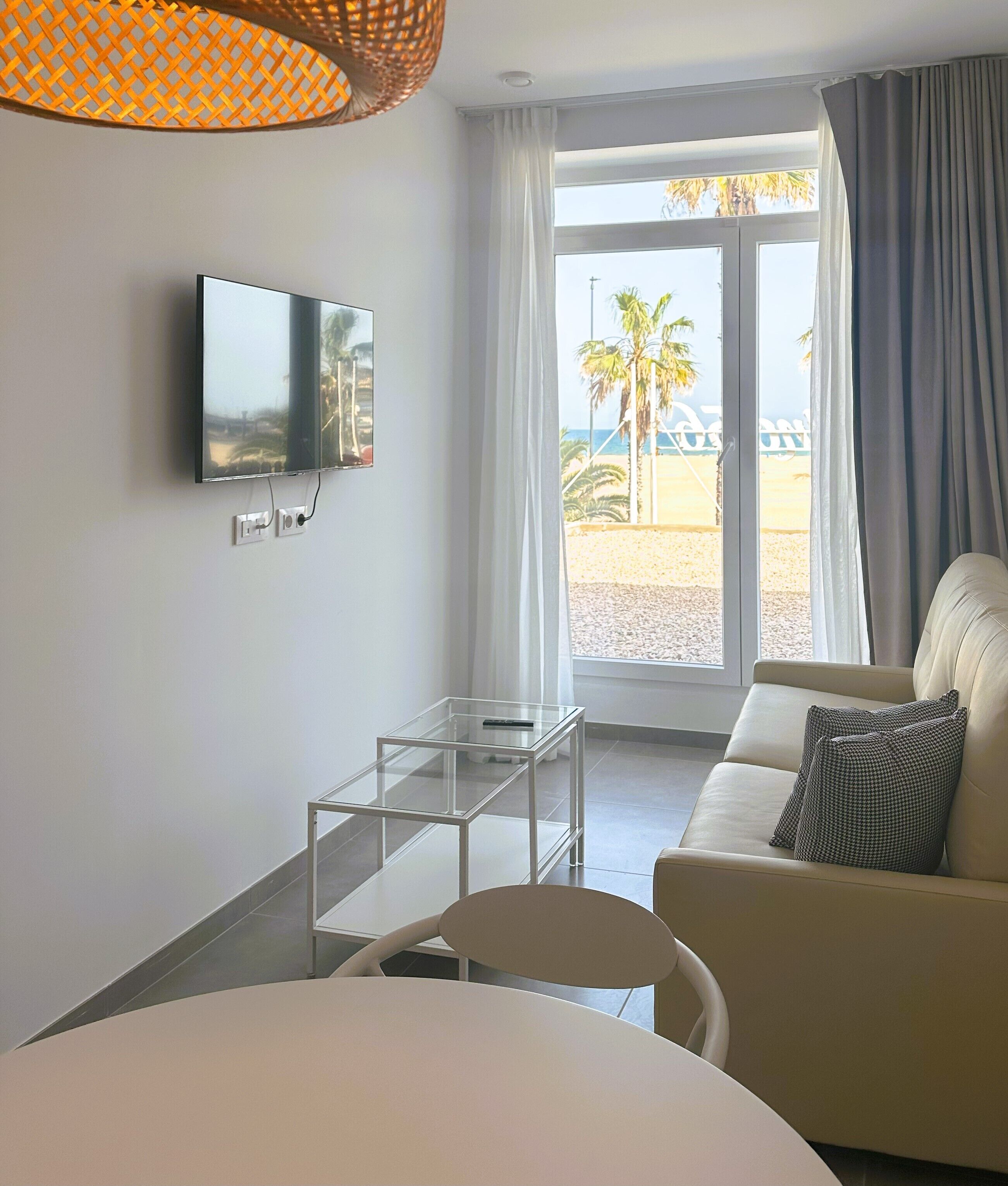 Een moderne hotelkamer met een flatscreen televisie aan de muur, een glazen salontafel, een beige bank met gestreepte kussens, en uitzicht op het strand door grote ramen.
