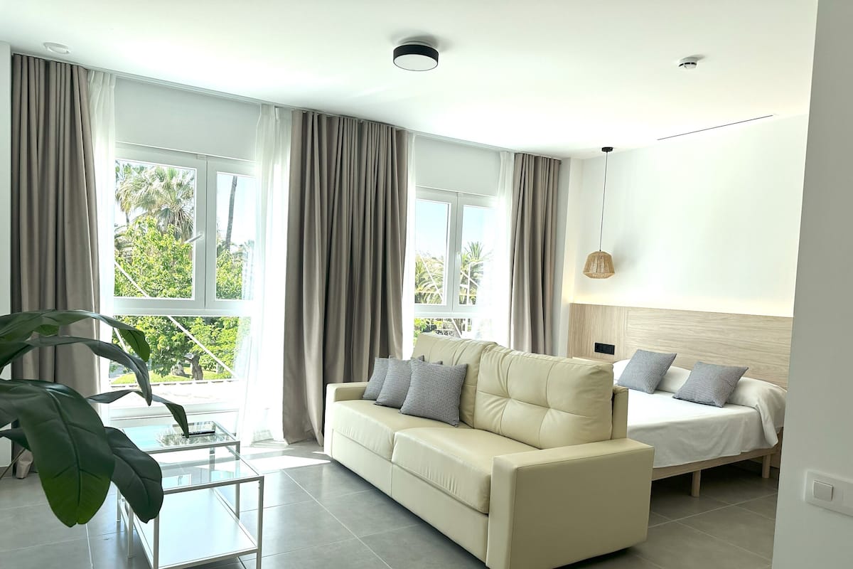 Un salon moderne avec un canapé beige, une table basse en verre et un lit doté d’une tête de lit en bois.