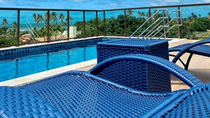 Uma área na piscina com cadeiras de vime azuis, céu azul e palmeiras ao fundo.