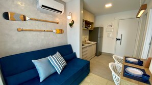 Uma sala de estar moderna com um sofá azul, uma mesa de jantar com pratos e um controle remoto, e uma cozinha com armários e eletrodomésticos.