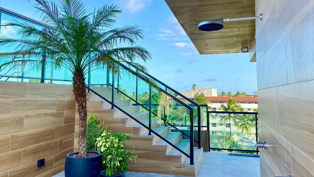 Uma escadaria moderna com guarda-corpos de vidro, que leva a uma varanda com vista para palmeiras e edifícios.