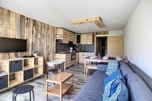 A modern living space with a wooden wall, a flat-screen TV, a sofa, a dining table, and a kitchen area. - 8th floor, balcony, tv, ski locker, 40m², Les Menuires (Les Menuires)
