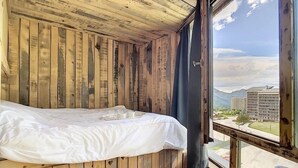 A wooden cabin room with a bed, a window, and a view of mountains and buildings. - Ski-in/ski-out & foot of the slopes, 8th floor, balcony, tv, ski locker, 40m², Les Menuires (Les Menuires)