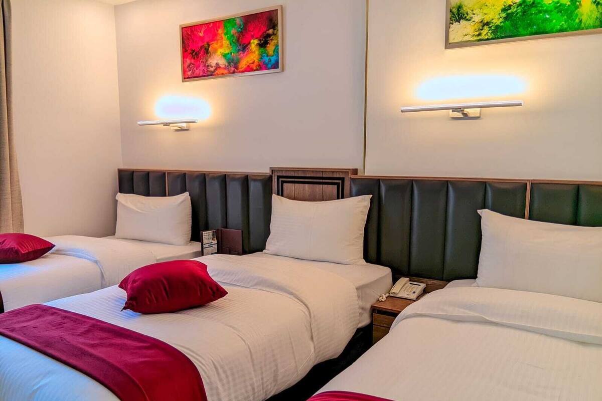 Habitación de hotel con dos camas, cada una con ropa blanca y almohadas rojas, una mesita de noche con un teléfono y un cuadro abstracto colorido en la pared.