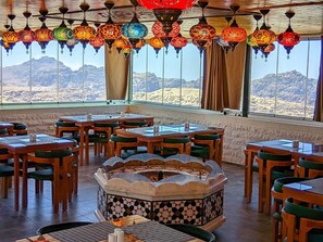 A restaurant with a central fire pit, multiple dining tables, and chairs. Large windows offer a view of the mountains. - Petra Bermudez Hotel  (Wadi Musa)