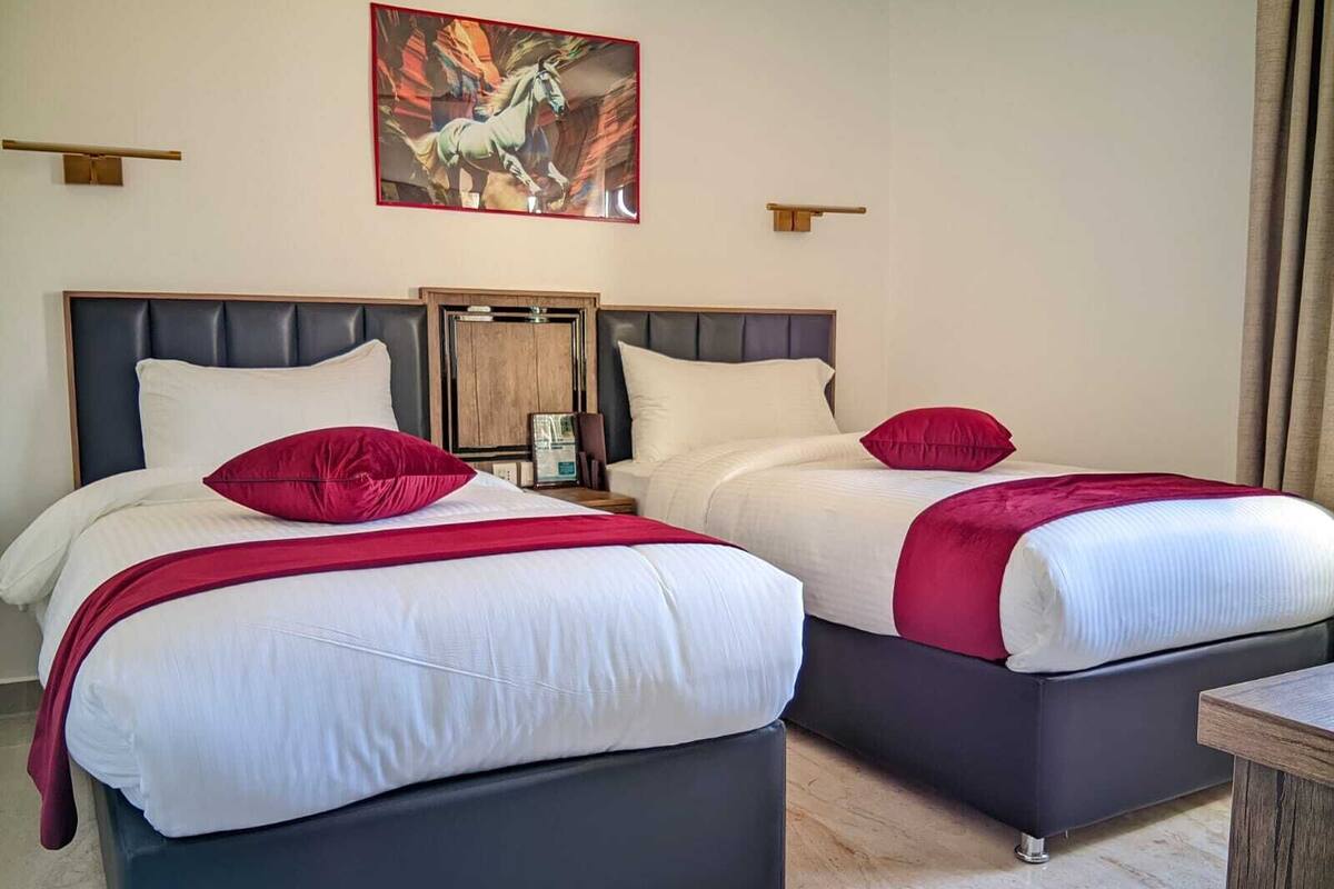 Habitación de hotel con dos camas, cada una con ropa blanca y almohadas rojas. Se observa un cabecero de madera, una mesita de noche y un cuadro de un caballo en la pared.