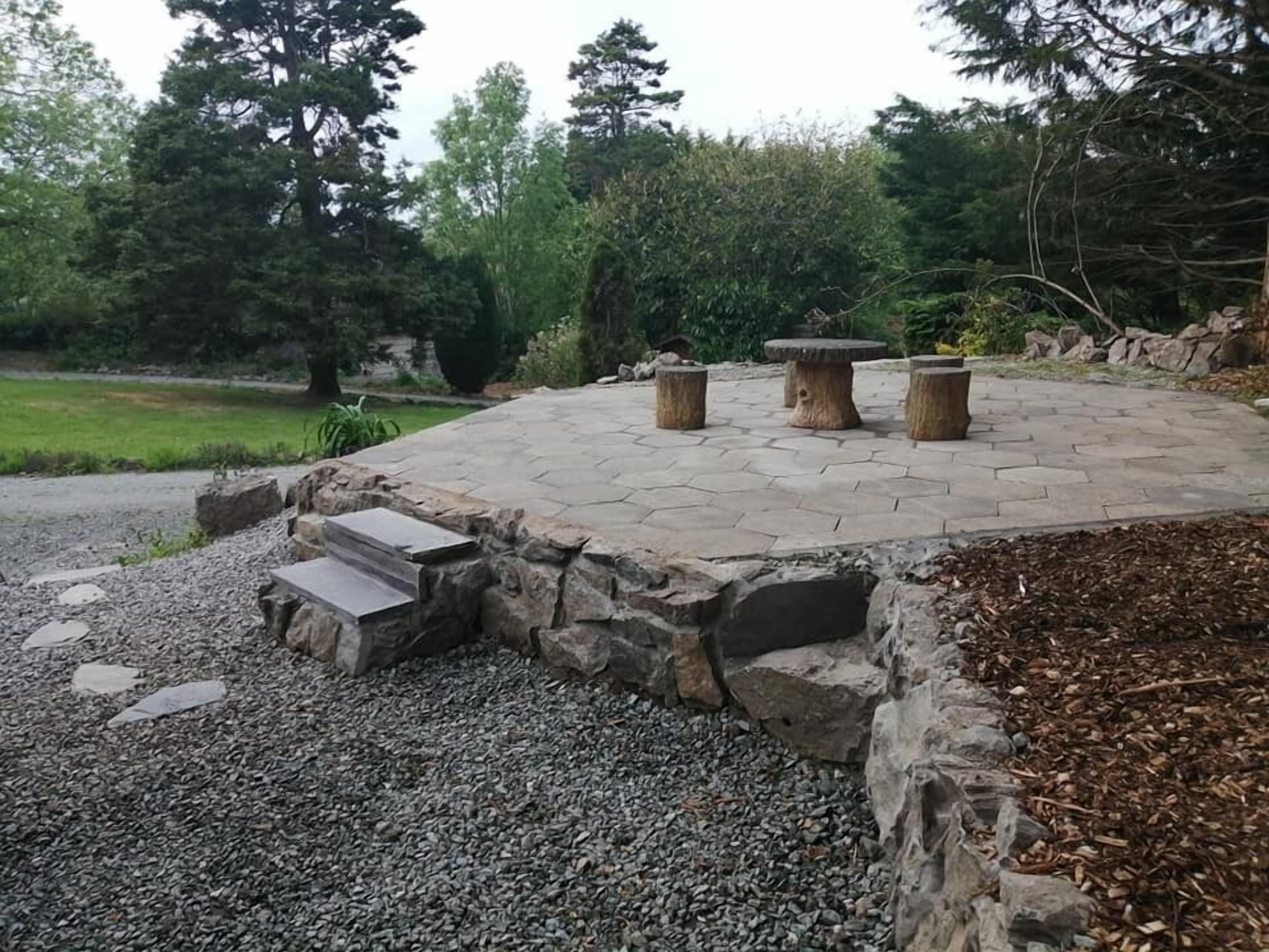 A stone patio with a seating area, surrounded by a stone wall and steps leading down to a gravel path.