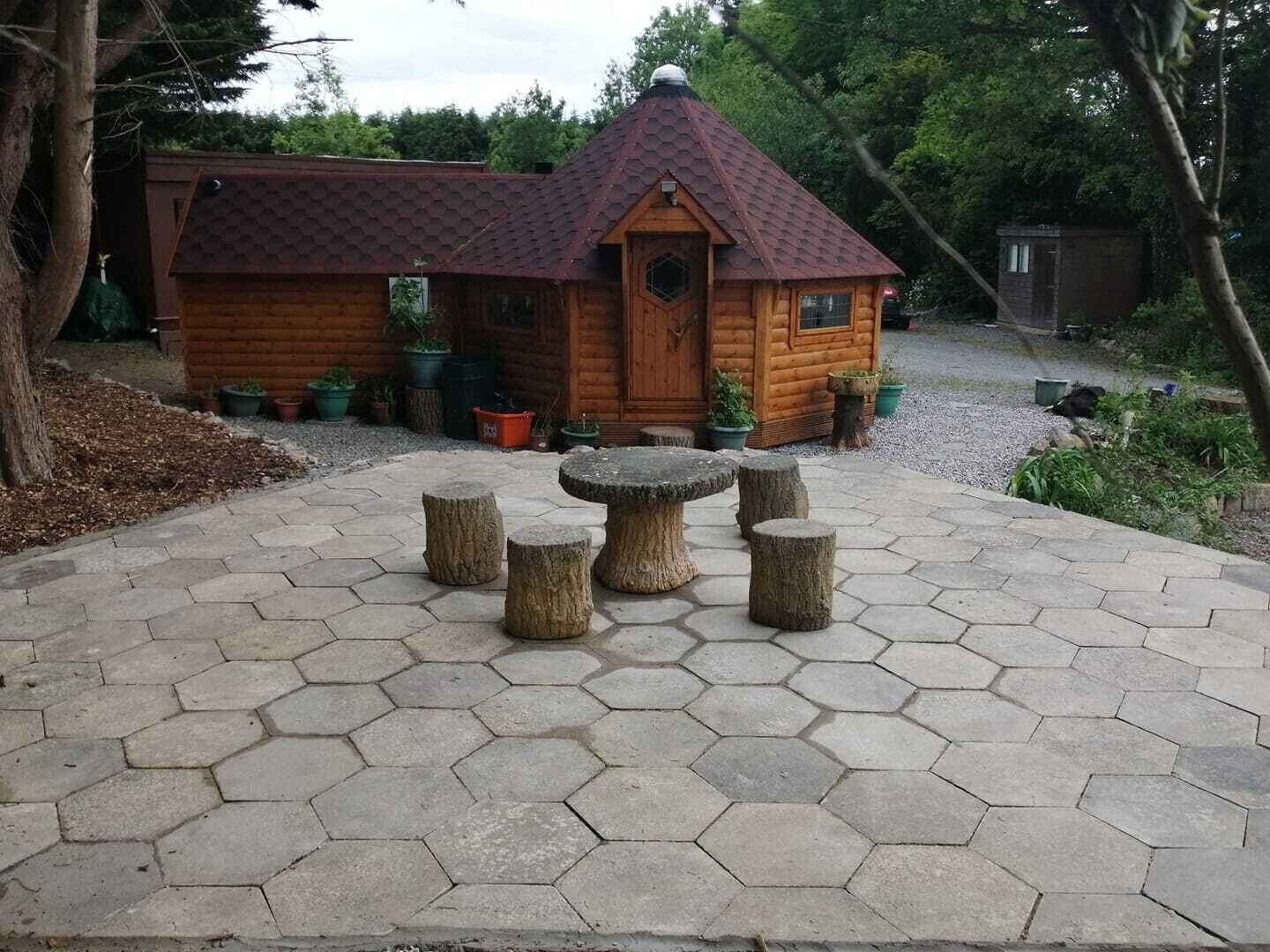A wooden cabin with a hexagonal tiled patio and a central stone table surrounded by stools.