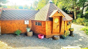 Een klein houten huisje met een zeshoekig betegeld dak, een houten deur met een ruit en een grindpad.