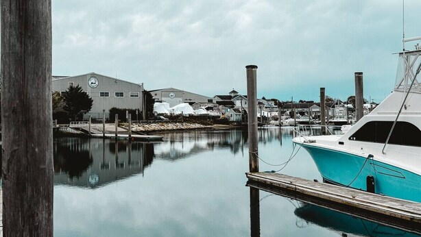 A marina with a docked boat, a large industrial building, and a calm water body reflecting the surroundings.