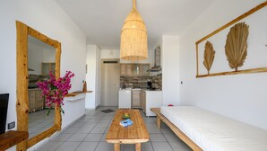A compact living space with a wooden table, a sofa, a hanging light fixture, a mirror, and a kitchen area. - Kalimera apartment 5 ,Near beach and all amenities (Adelianos Kampos)