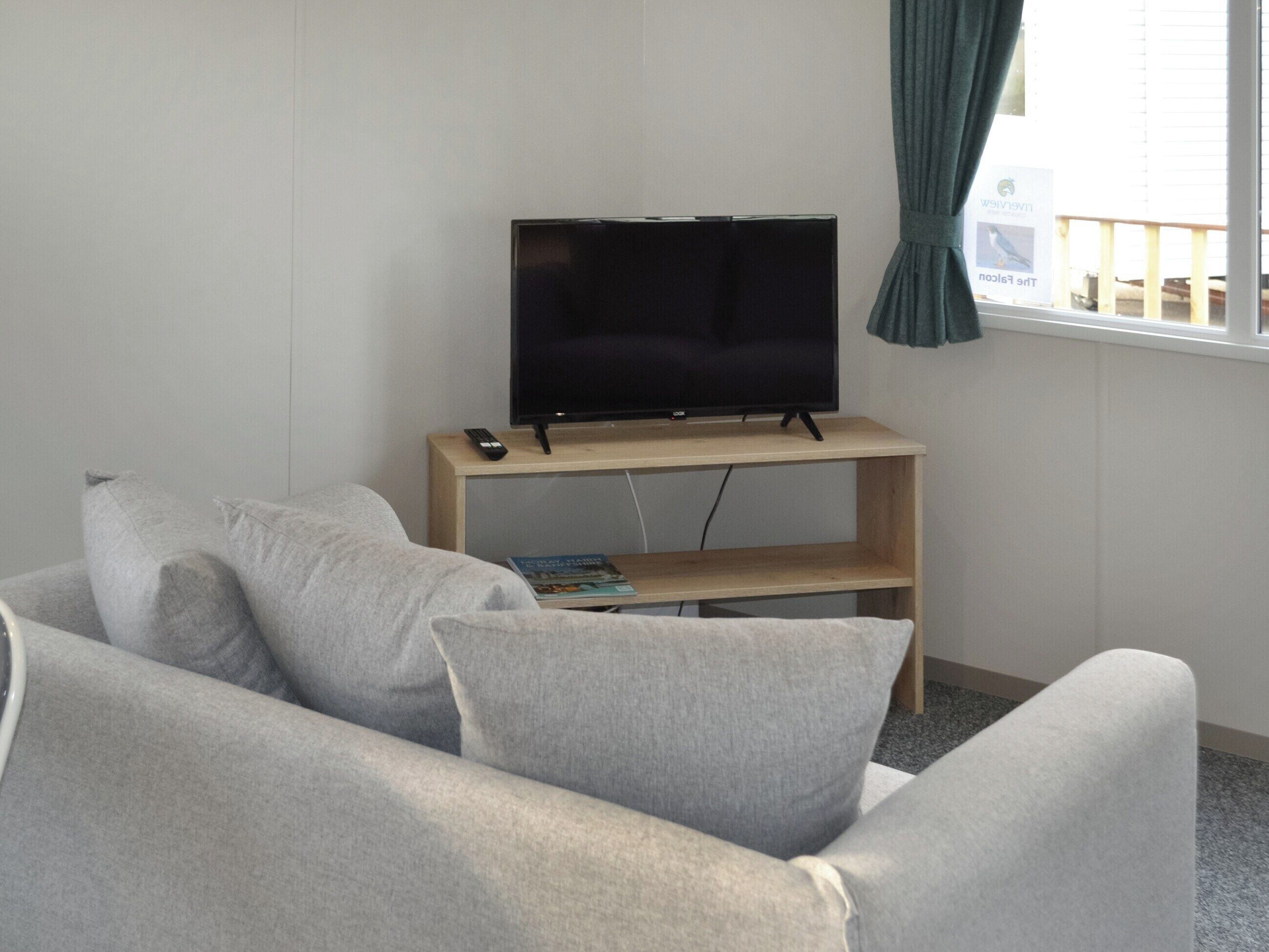 A living room with a flat-screen TV on a wooden stand, grey sofas, and a window with curtains.