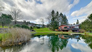 A house with a pond in front, surrounded by trees and a grassy area.