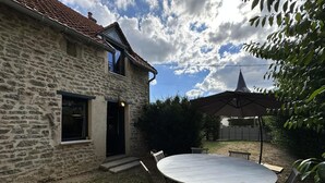 A stone house with a tiled roof, a round patio table with chairs, and a covered outdoor area. - Gite Climatisé les Lauriers (Essey)