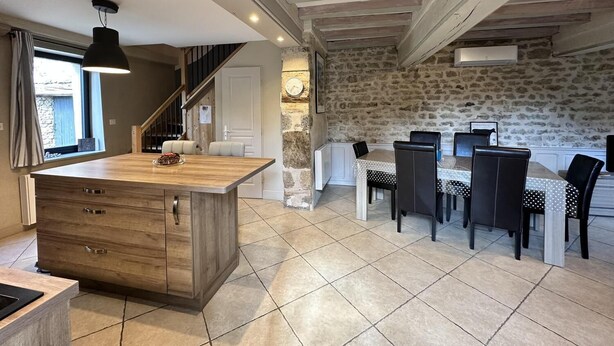 A modern kitchen with a central island, dining area with chairs, a staircase, and a stone wall. - Gite Climatisé les Lauriers (Essey)