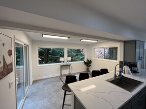 Una cucina moderna con un piano di lavoro in marmo, sedie nere, un tavolo da pranzo e ampie finestre che si affacciano su alberi.