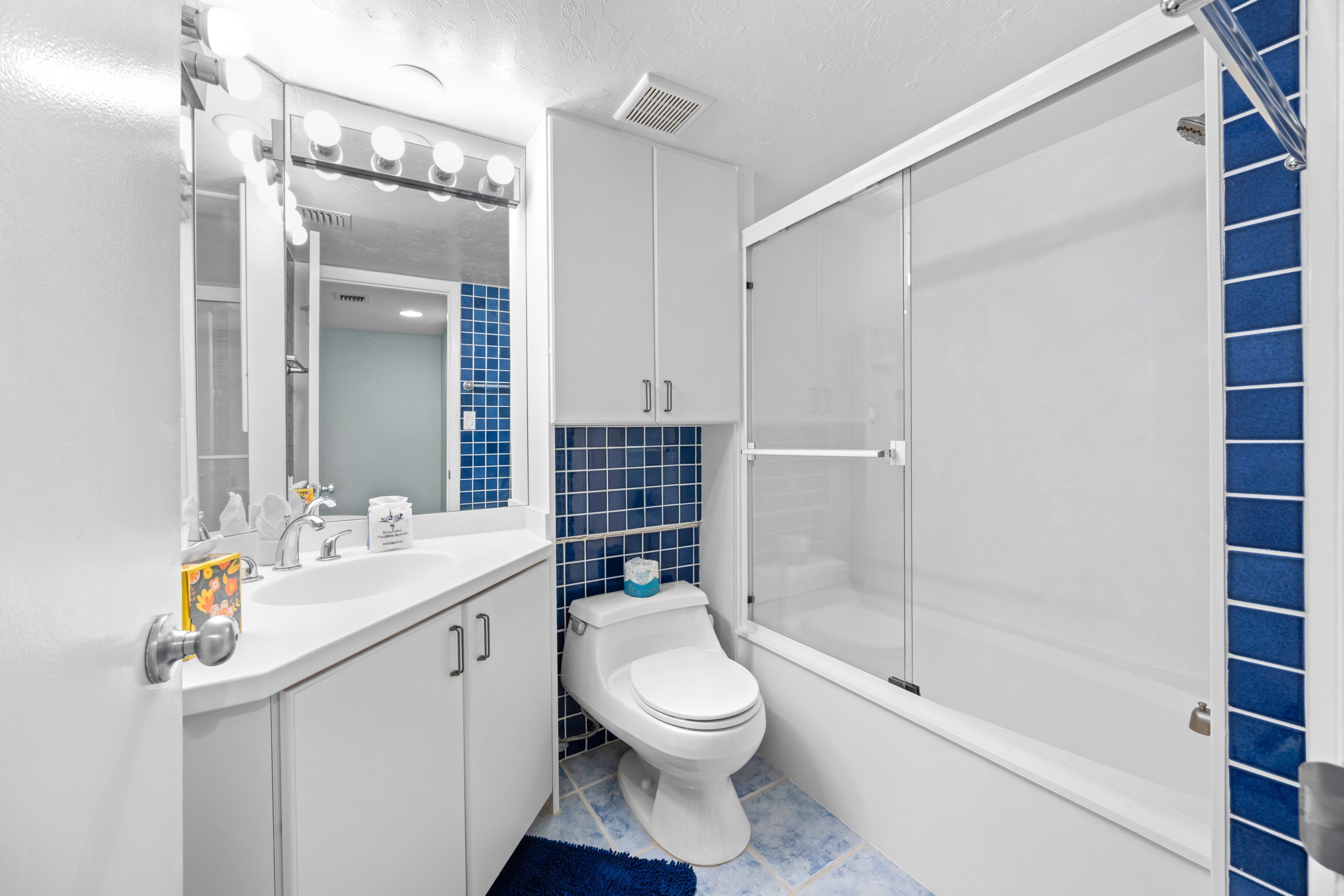 A bathroom with a white sink, mirror, and toilet, a blue tiled shower area, and a white bathtub.