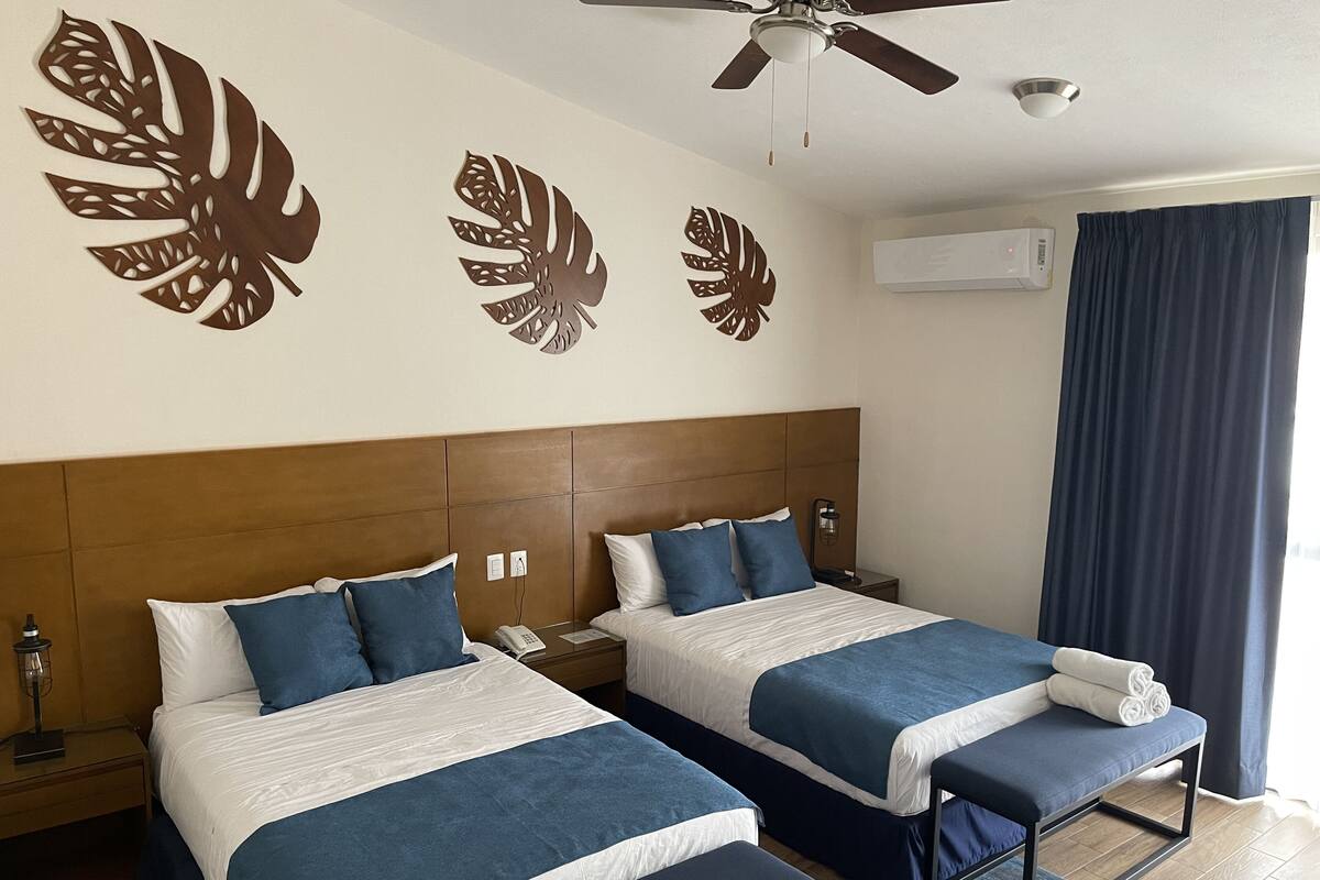 Habitación de hotel con dos camas, ventilador de techo, aire acondicionado y un ventanal con cortinas.