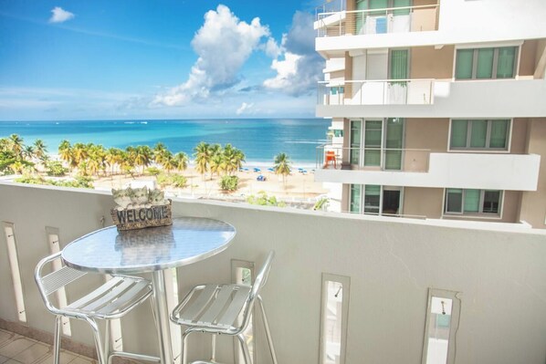 A balcony with a round table and two chairs, overlooking a beach and ocean.