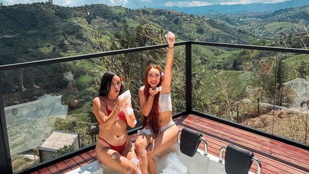 Dos personas en un jacuzzi en un balcón con una vista panorámica de un valle y montañas.