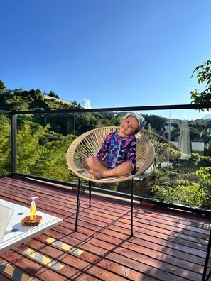 A person seated in a wicker chair on a wooden deck with a glass balustrade, overlooking a lush landscape. - Luum Glamping by Roots Rentals (Marinilla)