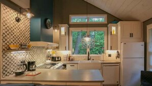 A kitchen with a central island, white cabinets, a window with a view of greenery, and a hanging light fixture. - Updated cottage "Beriozka" on Cedar Lake (Chester)