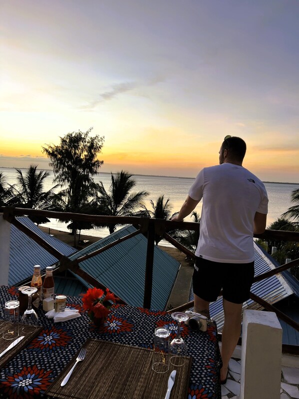 Un homme, debout sur un balcon, contemple le coucher du soleil. Une table est dressée pour deux, avec une nappe à motifs et une bouteille de vin.