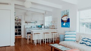 Une cuisine moderne avec des meubles blancs, un îlot central doté de tabourets et un coin repas comprenant un canapé et des coussins.