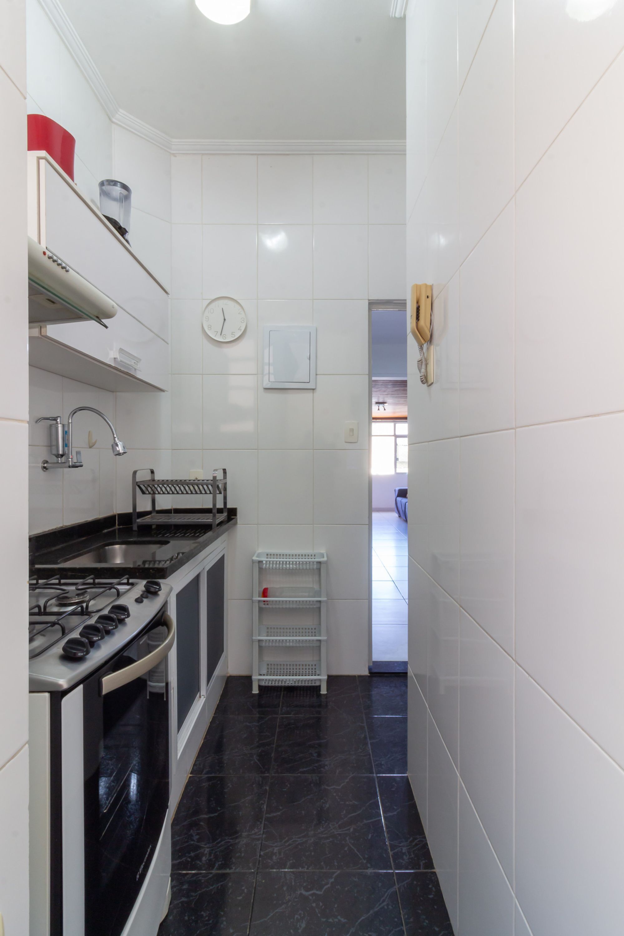 Een smalle keuken met witte tegels, een fornuis, een spoelbak en een klok aan de muur.