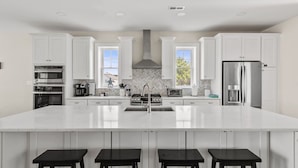 A modern kitchen with white cabinetry, a large island with bar stools, stainless steel appliances, and a herringbone tile backsplash. - Fenwick on Bay mins to beach. Hot tub, Game Rm! (Selbyville)