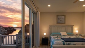 A hotel room with a bed, bedside lamps, a painting, and a view of a sunset over a waterfront. - Fenwick on Bay mins to beach. Hot tub, Game Rm! (Selbyville)