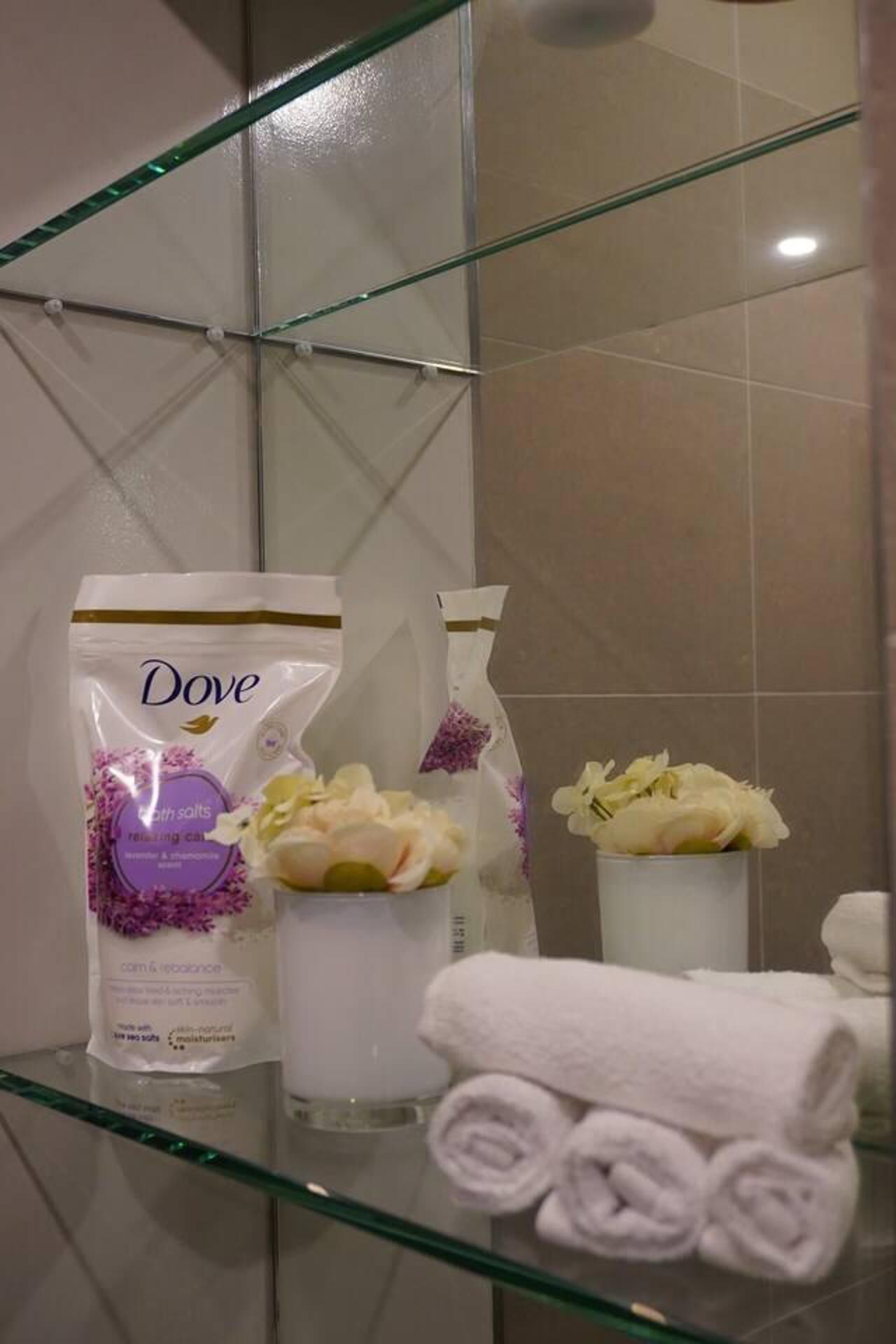 A bathroom with a glass shower enclosure, a Dove soap bar, a bottle of Dove lotion, and a rolled white towel on a glass shelf.