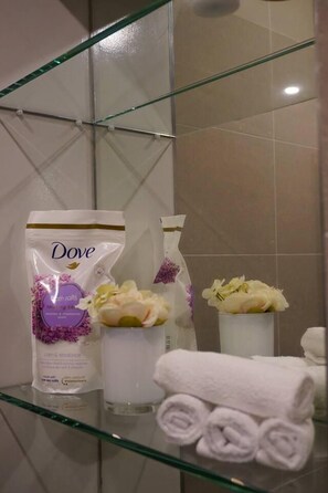 A bathroom with a glass shower enclosure, a Dove soap bar, a bottle of Dove lotion, and a rolled white towel on a glass shelf. - The Glamour of Shoreditch (London)
