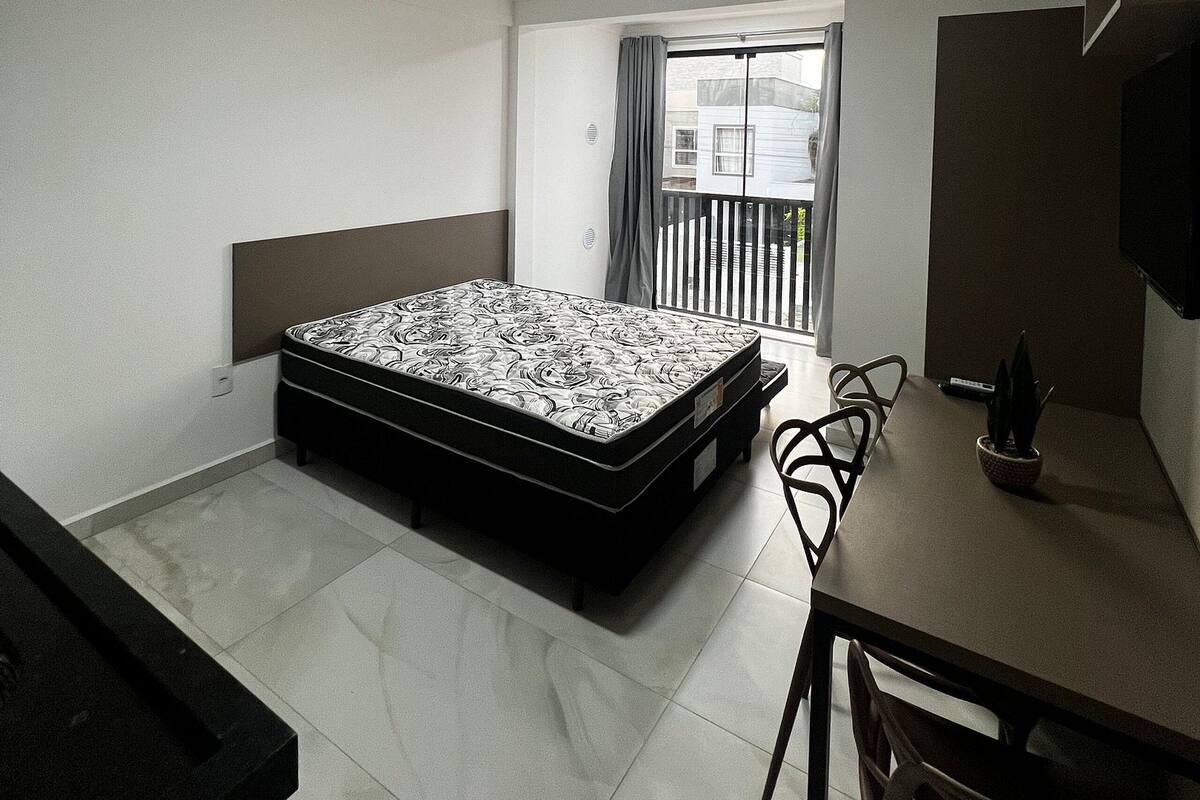 A small, clean room with a bed, a desk, and a chair. There is a window with curtains and a view of the outside.
