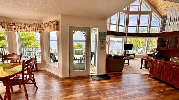 A spacious living area with a wooden floor, a dining table with chairs, a sofa, and a TV. Large windows provide views of the outdoors. - Azure Ocean Beach House (Port McNeill)
