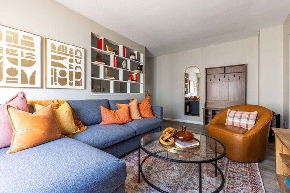 A living room with a blue sofa, orange cushions, a glass coffee table, and a wooden cabinet.