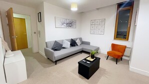 A modern living room with a grey sofa, a black coffee table, and an orange armchair. There is a door leading to another room, a window with a view, and a ceiling light. - Central 2-bed Apartment in Greater Manchester (Manchester)