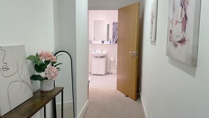 A hallway with a wooden door leading to a bathroom. There is a mirror on the left wall, a painting, and a vase with flowers on a console table. - Central 2-bed Apartment in Greater Manchester (Manchester)