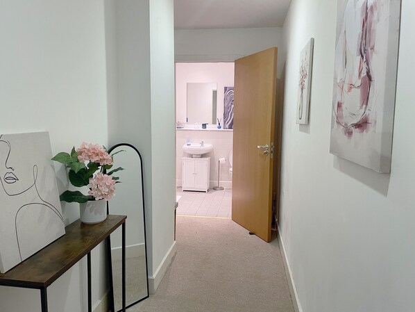A hallway with a wooden door leading to a bathroom. There is a mirror on the left wall, a painting, and a vase with flowers on a console table.