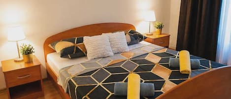 A neatly made bed with a geometric patterned bedspread, two bedside tables with lamps, and a potted plant on one of the tables.