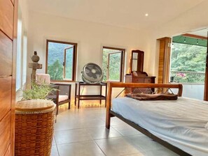 A bedroom with a bed, a fan, a dresser, and a window with a view of greenery.