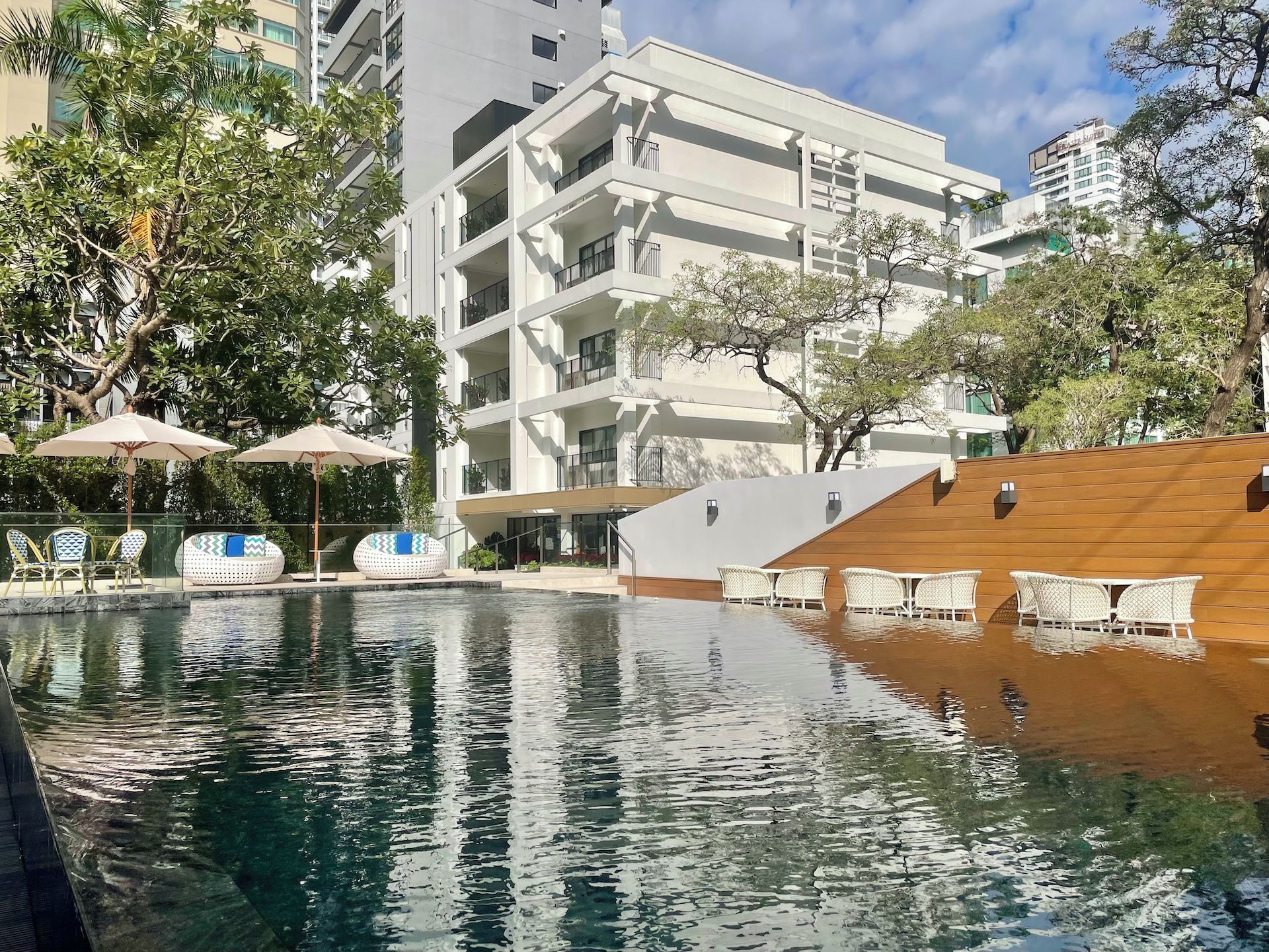 Una piscina con sillas de descanso y sombrillas, rodeada de edificios de varios pisos y árboles.