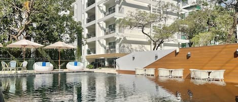 A swimming pool with lounge chairs and umbrellas, surrounded by multi-story buildings and trees.