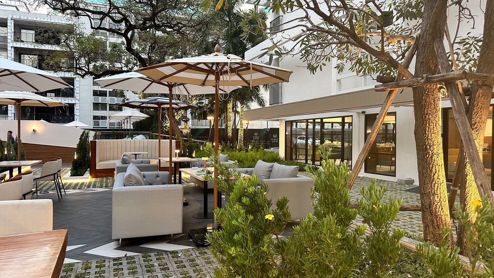 An outdoor seating area with modern furniture and umbrellas, surrounded by greenery and a building with large windows.