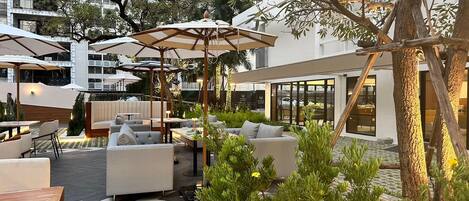 Un espace extérieur aménagé avec des meubles modernes et des parasols, entouré de verdure et bordé d’un bâtiment doté de grandes fenêtres.