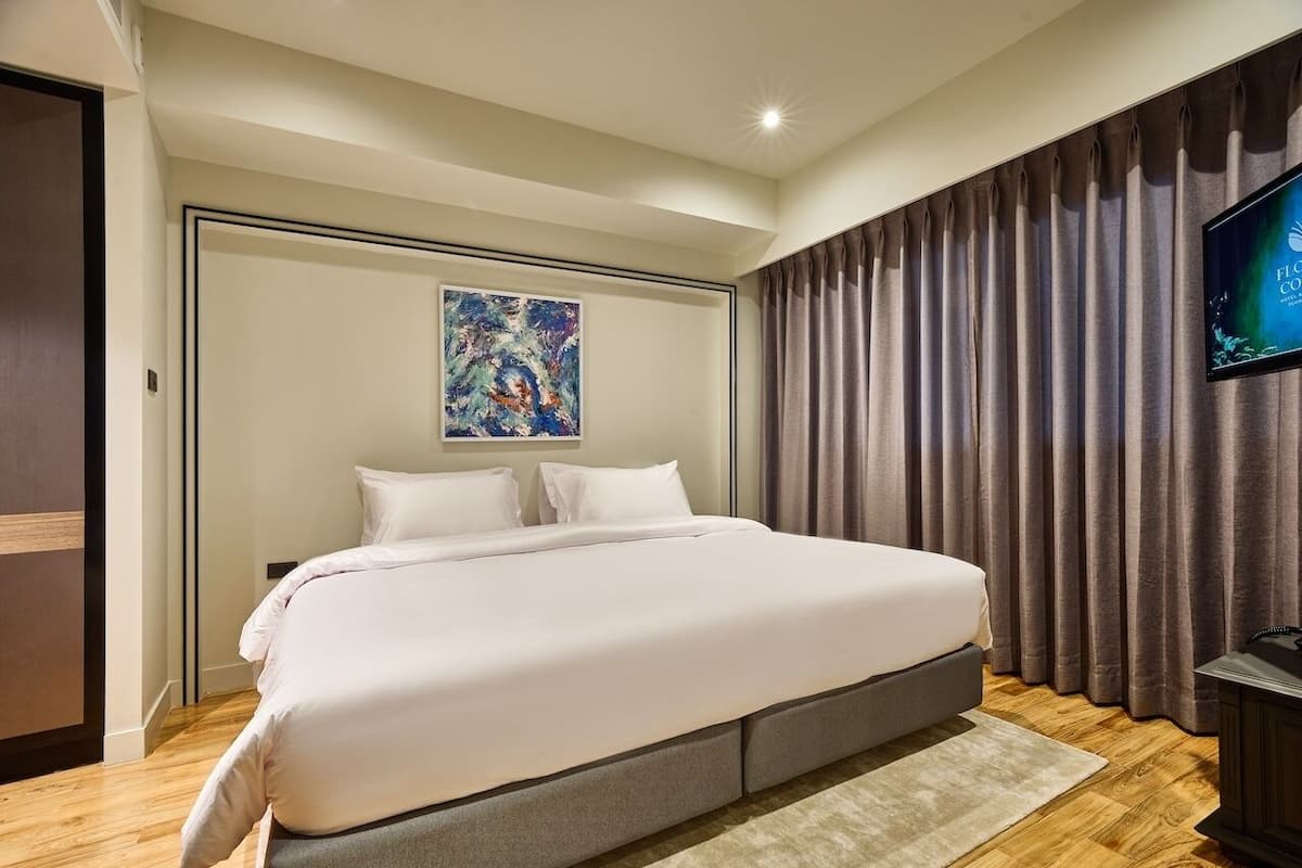 A neatly made bed in a hotel room with a large painting above it, a television on the wall, and a wooden floor.