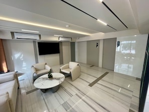 A modern hotel room with a flat-screen TV, two armchairs, a round coffee table, and a large mirror. - Floral Court Hotel and Residence (Bangkok)